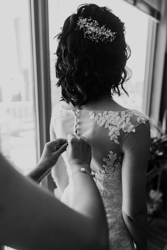 Black and white photo of woman buttoning bride's dress as they stand by a window before Ada Street Chicago wedding celebration