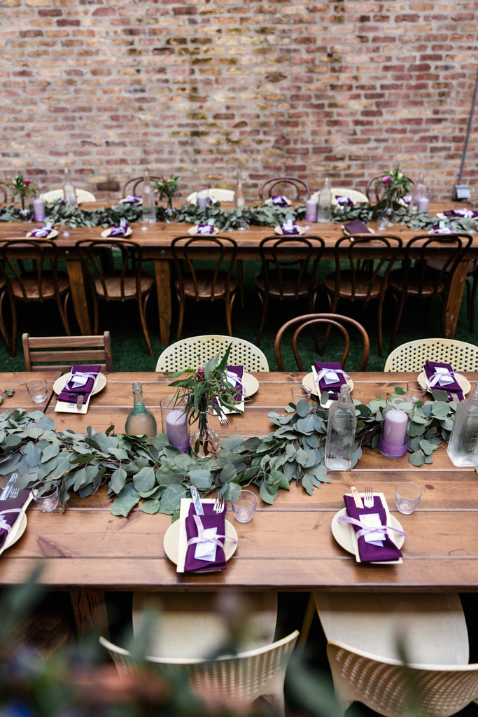 Guest tables set for a meal with greenery, purple napkins, and candles against a brick wall for wedding reception at Ada Street Chicago
