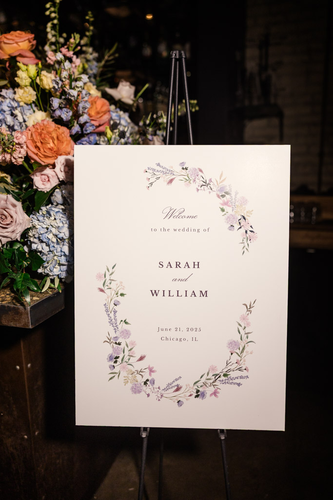 Wedding welcome sign with a floral border, displayed next to an arrangement of flowers at Ada Street Chicago wedding celebration