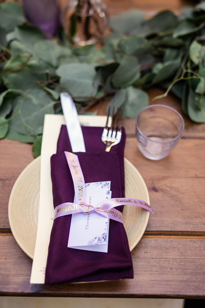 Guest place setting for Ada Street Chicago wedding reception features a purple napkin, menu, knife, fork, and a glass on a wooden table adorned with green leaves décor