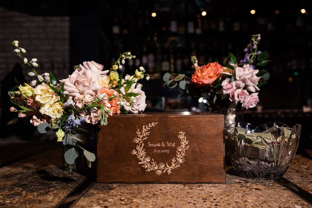 Wooden wedding card box with engraved names, surrounded by floral arrangements and a glass bowl full of commemorative matchboxes for wedding celebration at Ada Street Chicago