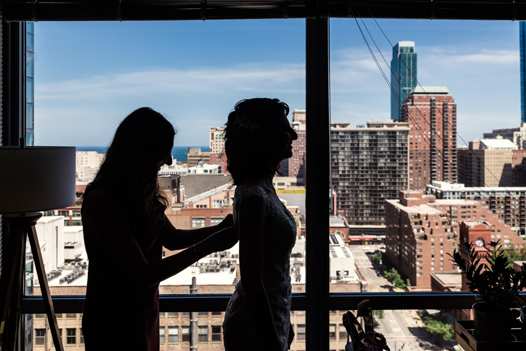 Two women in silhouette in front of a large window, one adjusting the bride’s dress, with Chicago skyscrapers in the background