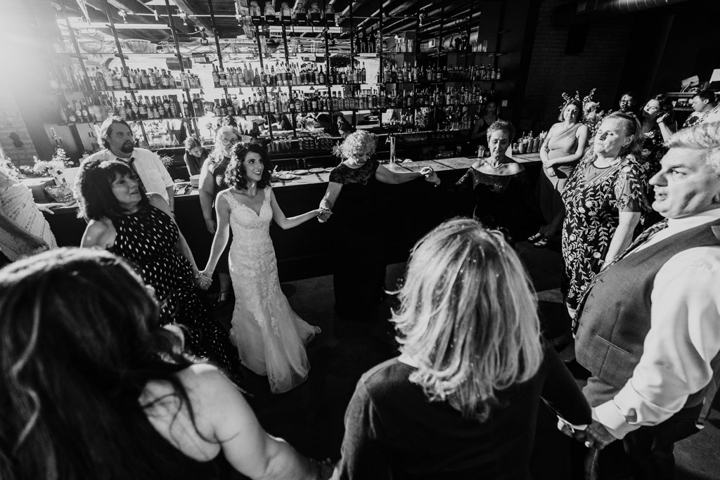 Black and white photo of bride and guests in formal attire stand in a circle, holding hands during wedding reception at Ada Street Chicago