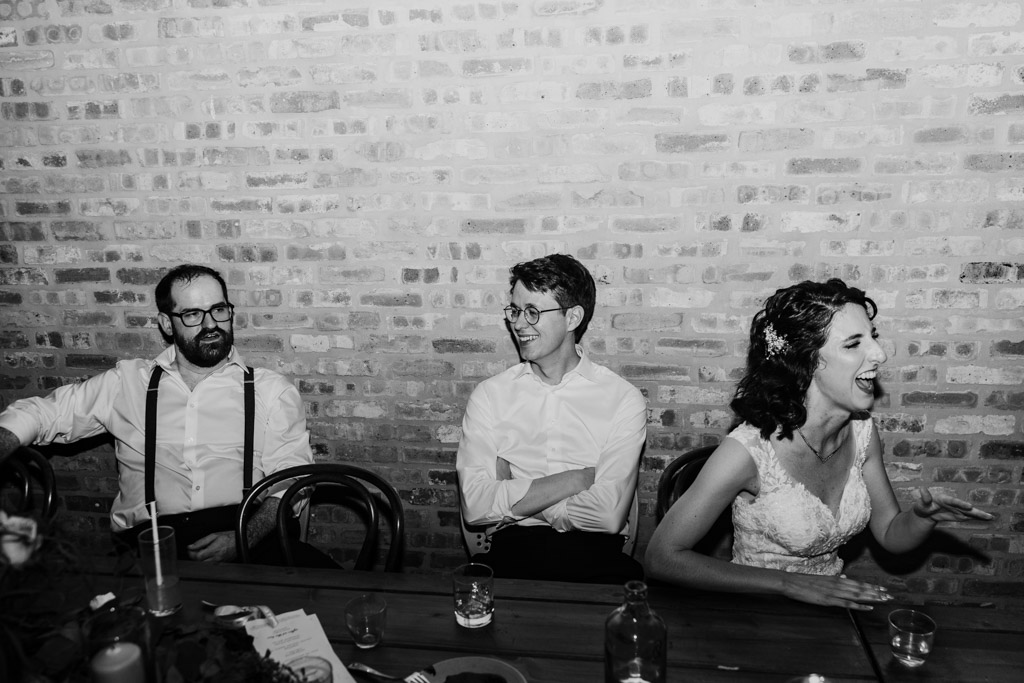 Black and white photo of bride, groom and Best Man sitting at a table laughing and talking in front of a brick wall at Ada Street Chicago