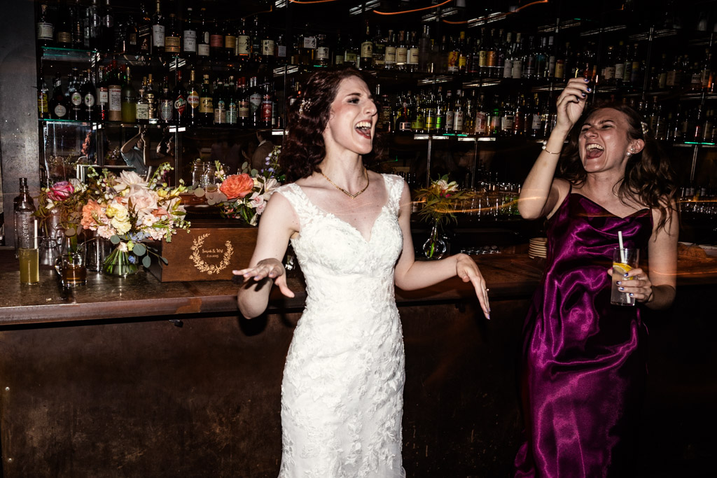 Bride and guest laugh and dance together at Ada Street Chicago, framed by a bar lined with bottles and vibrant floral arrangements