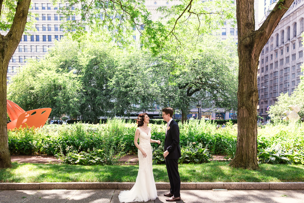 A bride and groom stand facing each other during their first look in a sunny, green Chicago park