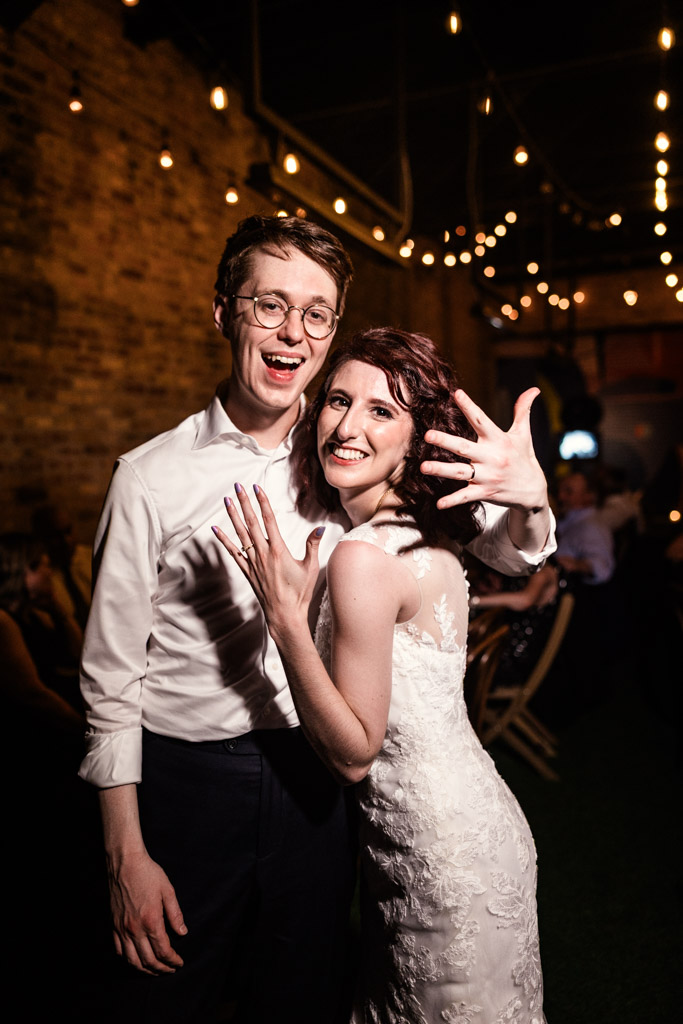 Smiling couple shows off their wedding rings at their nighttime wedding celebration under string lights at Ada Street Chicago
