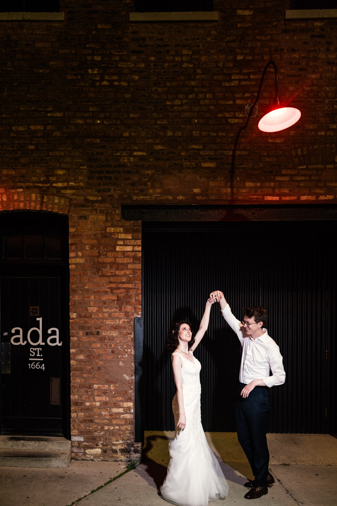 Bride and groom dance joyfully at night under a red light in front of Ada Street's brick facade