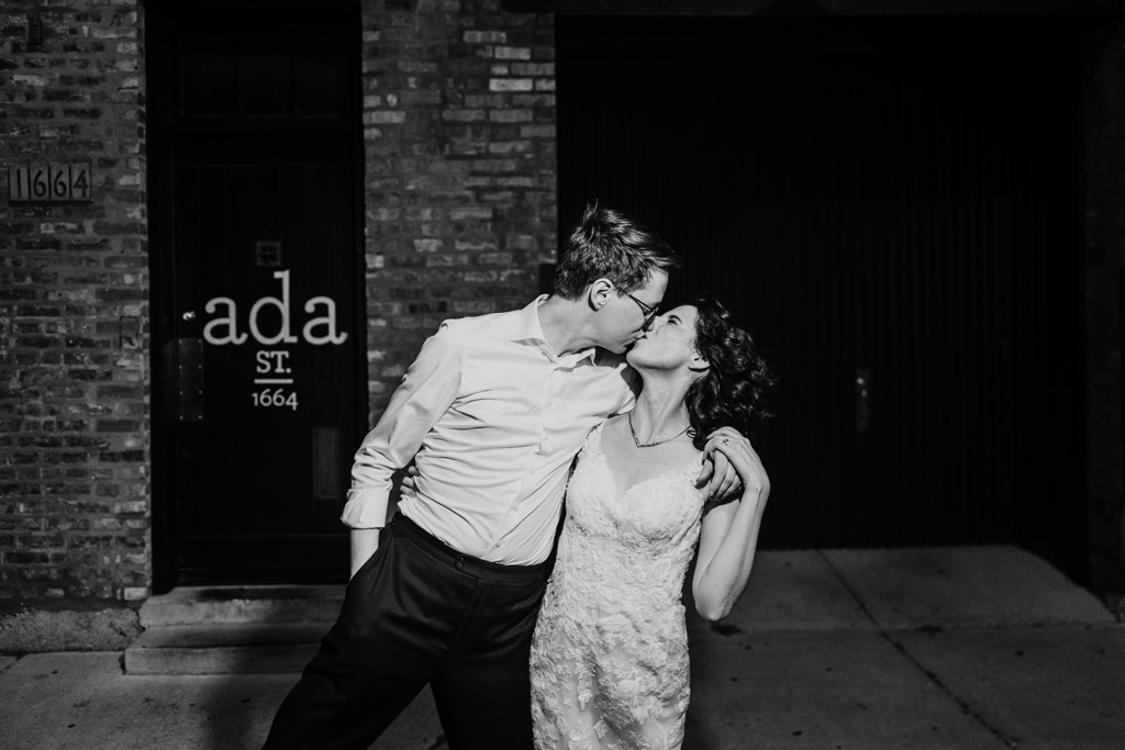Black and white photo of newlywed couple in wedding attire kissing outside Ada Street in Chicago at night
