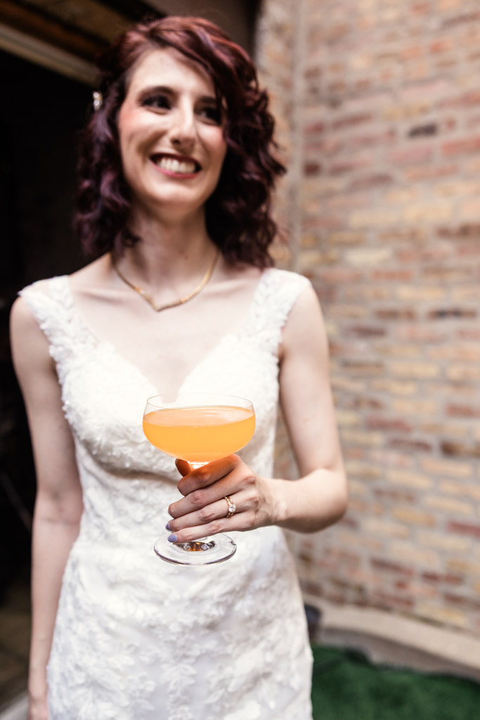 Bride in a white dress holds an orange custom cocktail in a coupe glass, standing by a brick wall inside Ada Street Chicago