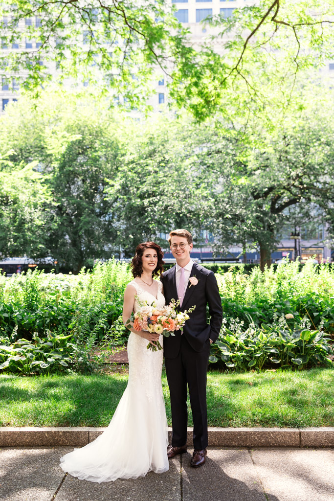 Portrait of bride and groom in a sunlit garden with lush greenery and tall trees in the background
