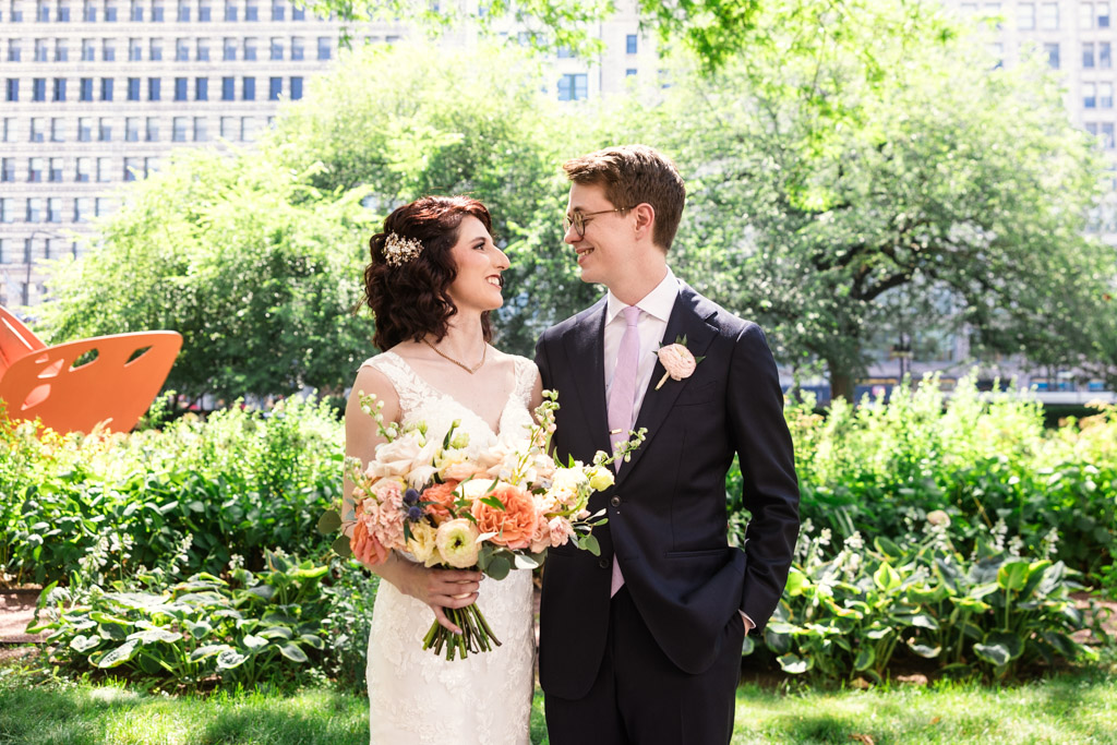 Bride and groom smiling at each other outside surrounded by greenery and flowers on a sunny day before their Ada Street wedding celebration