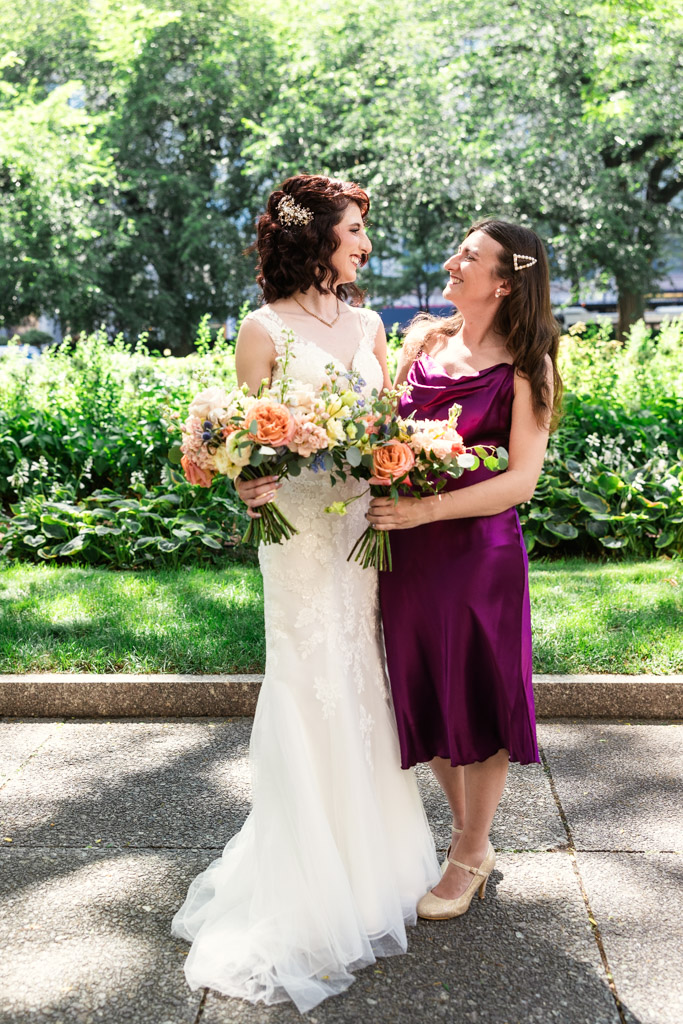 A bride in white and Maid of Honor in purple smile at each other, holding flower bouquets outdoors before Ada Street Chicago wedding celebration