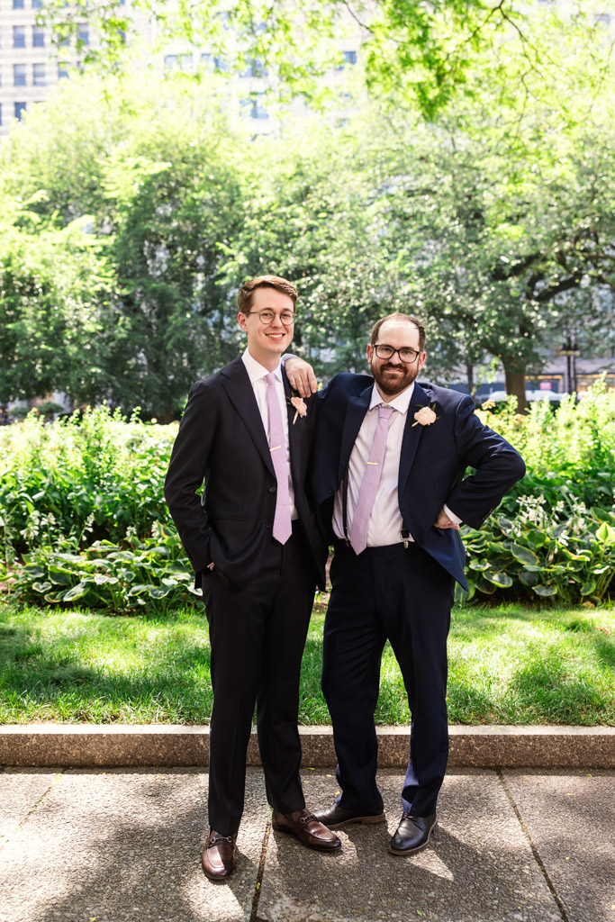 Casual portrait of groom and Best Man in suits with pink ties stand smiling together in a sunlit park with green trees and grass before Ada Street Chicago wedding celebration
