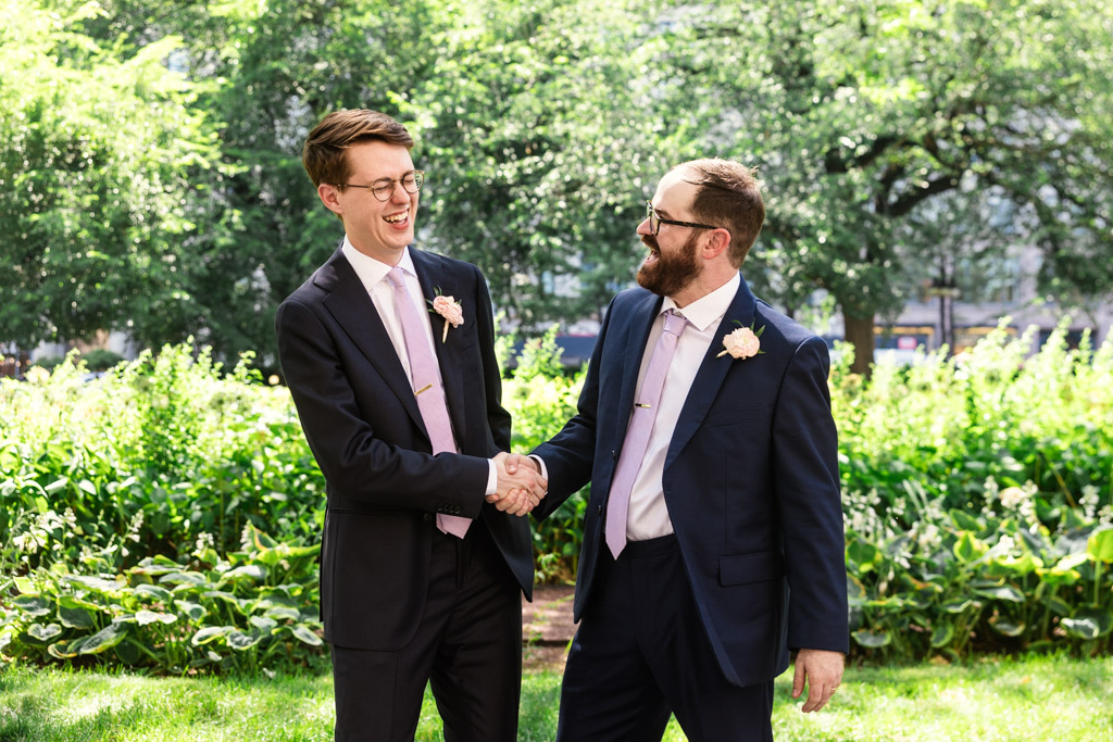 Groom and Best Man in suits and ties shake hands and smile at each other outdoors in a sunny, green park before wedding celebration at Ada Street Chicago