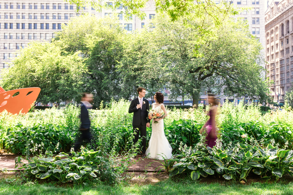 Creative photo of bride and groom standing together in a park as the Best Man and Maid of Honor walk toward them, motion-blurred