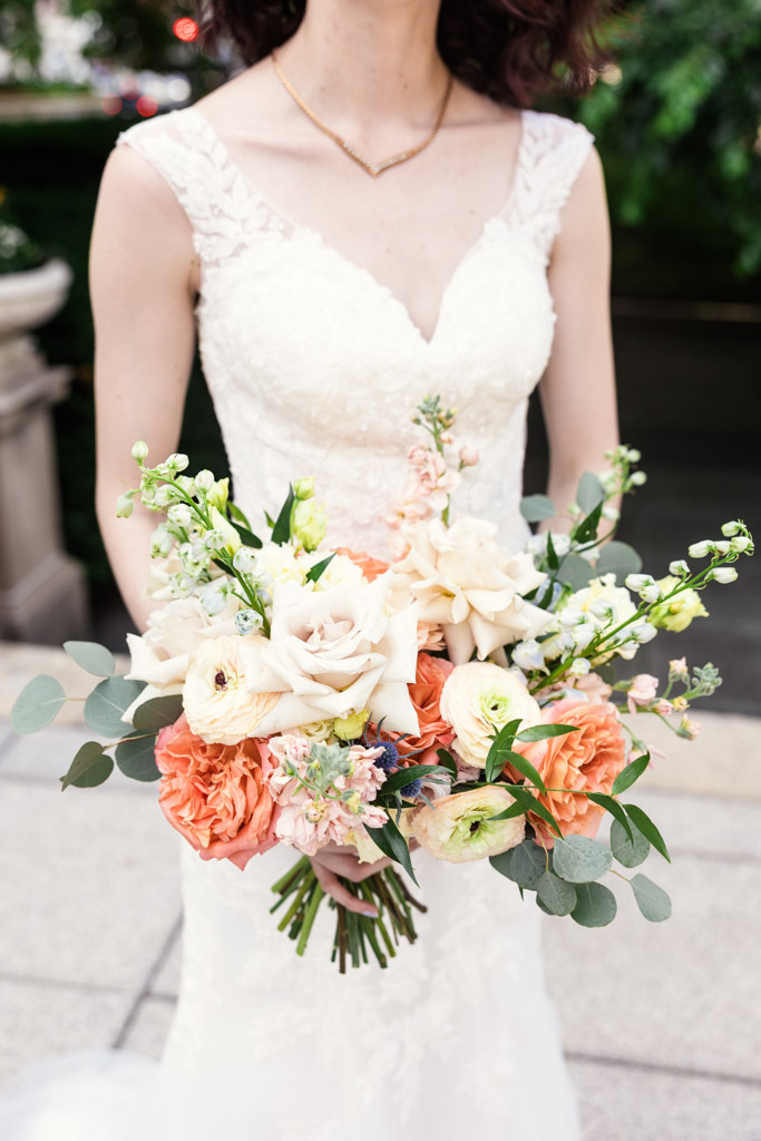 Bride in a white lace dress holding a bouquet of peach, white, and green flowers outdoors