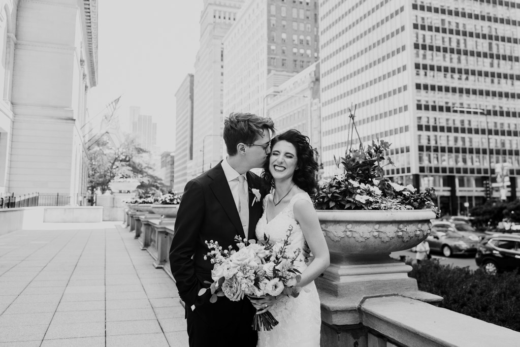 Black and white photo of bride and groom smiling, standing close together in front of Art Institute of Chicago with tall downtown buildings behind them