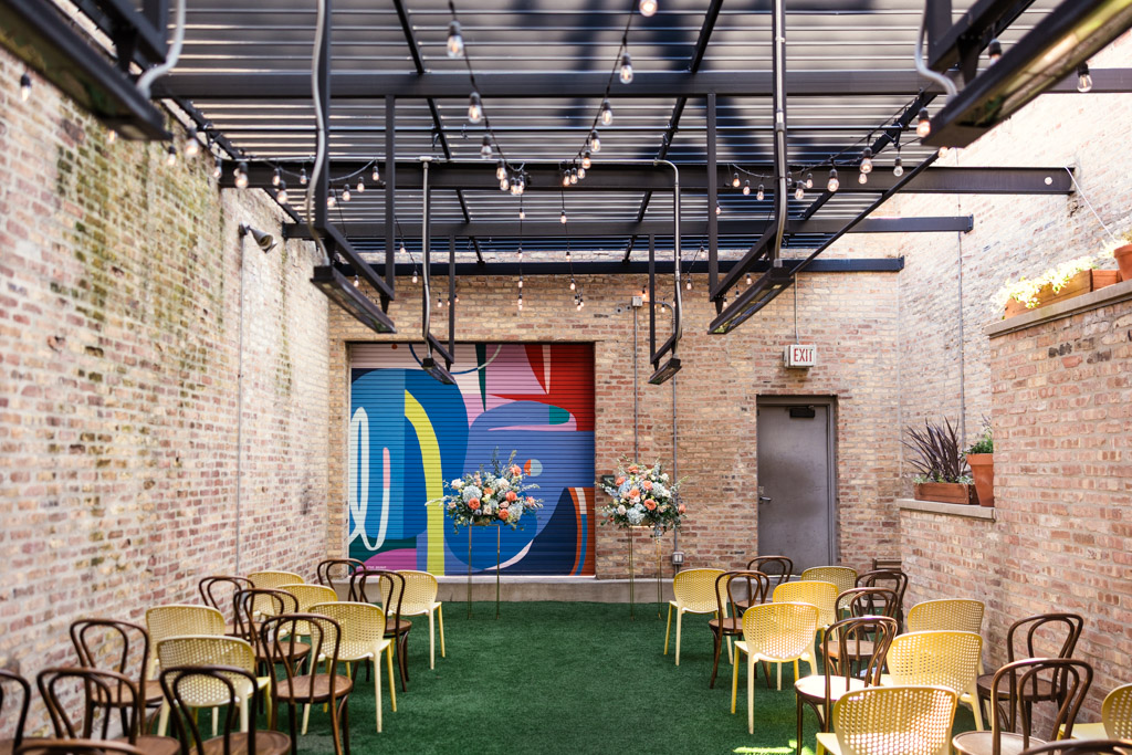Wedding ceremony space at Ada Street Chicago with yellow chairs, string lights, vibrant flower arrangements, and a colorful mural on a brick wall