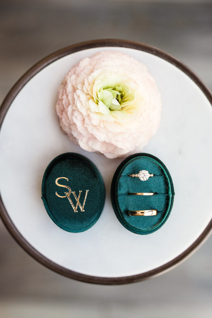 A flower and green velvet ring box with two rings and gold initials on a round white table for Ada Street Chicago wedding celebration