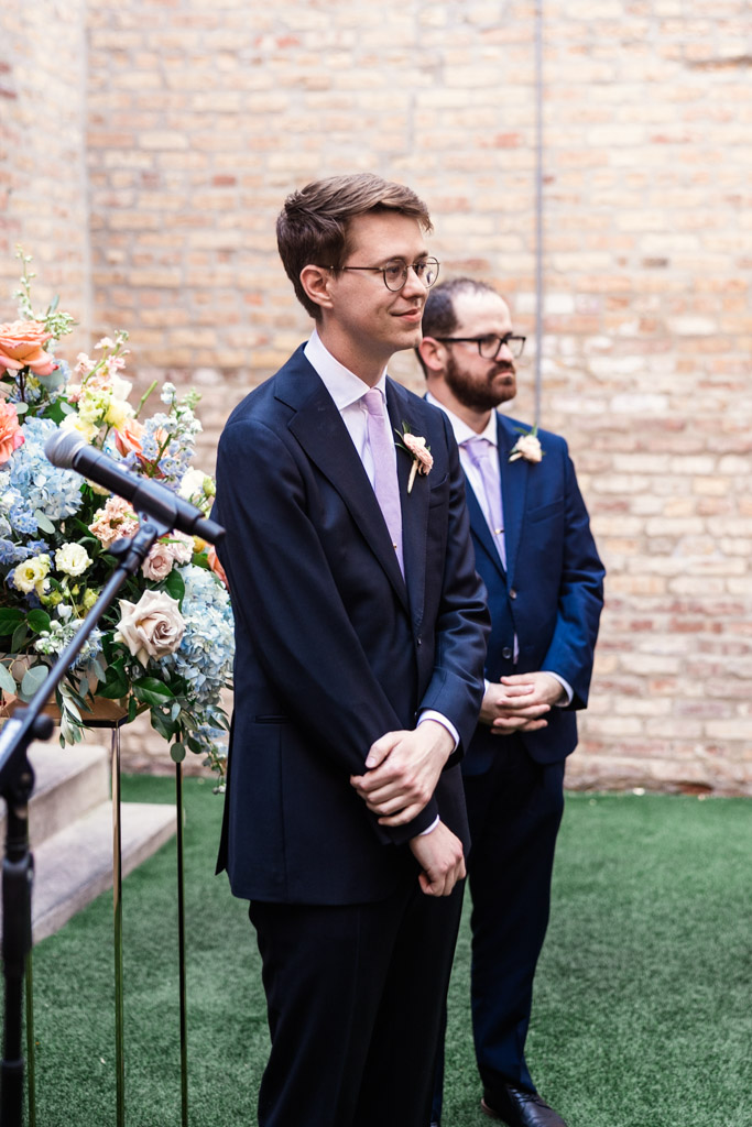 Groom watches bride walk down the aisle during wedding ceremony at Ada Street Chicago, with a brick wall in the background