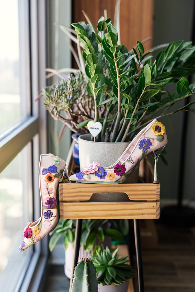Bride's pink high heels with colorful floral embroidery displayed on a wooden stand beside potted plants by a window for Ada Street Chicago wedding