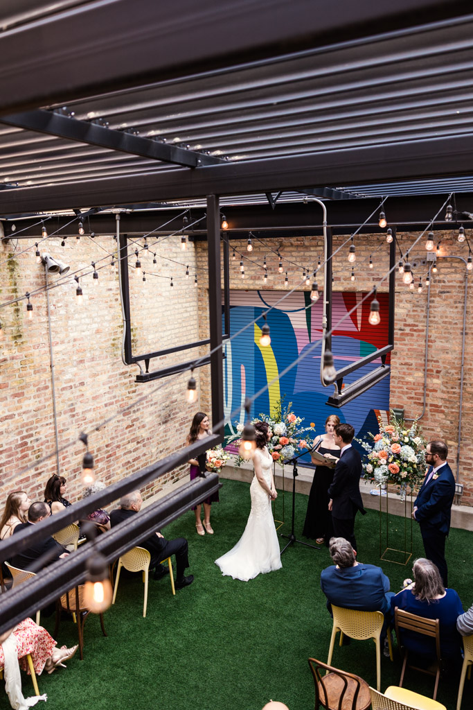 A wedding ceremony inside Ada Street Chicago, with guests seated, string lights overhead, and a colorful mural in the background