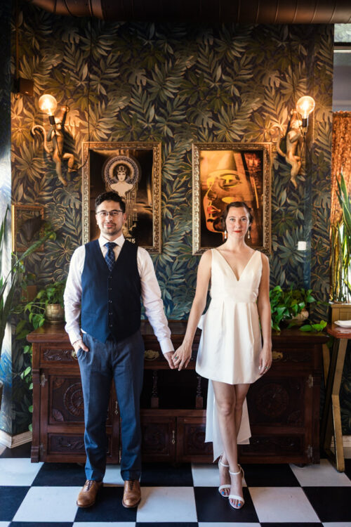 Creative portrait of engaged couple holding hands inside Cafe Deko in Chicago with vintage decor and patterened wallpaper behind them