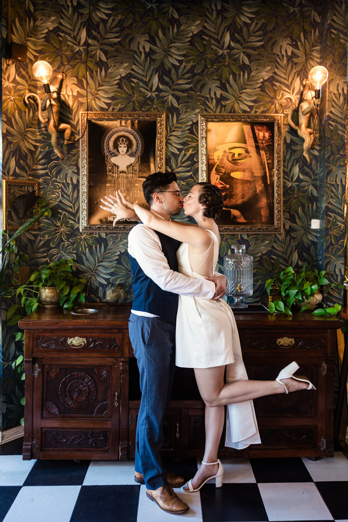 A couple in formal attire reminiscent of the 1920s embrace and kiss inside Cafe Deko, surrounded by patterned wallpaper and framed art