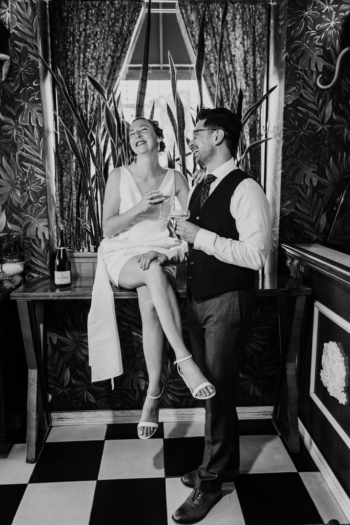 Black and white photo of happy couple in formal attire sharing drinks with the woman sitting on a counter in a stylish, plant-filled room during Cafe Deko engagement session