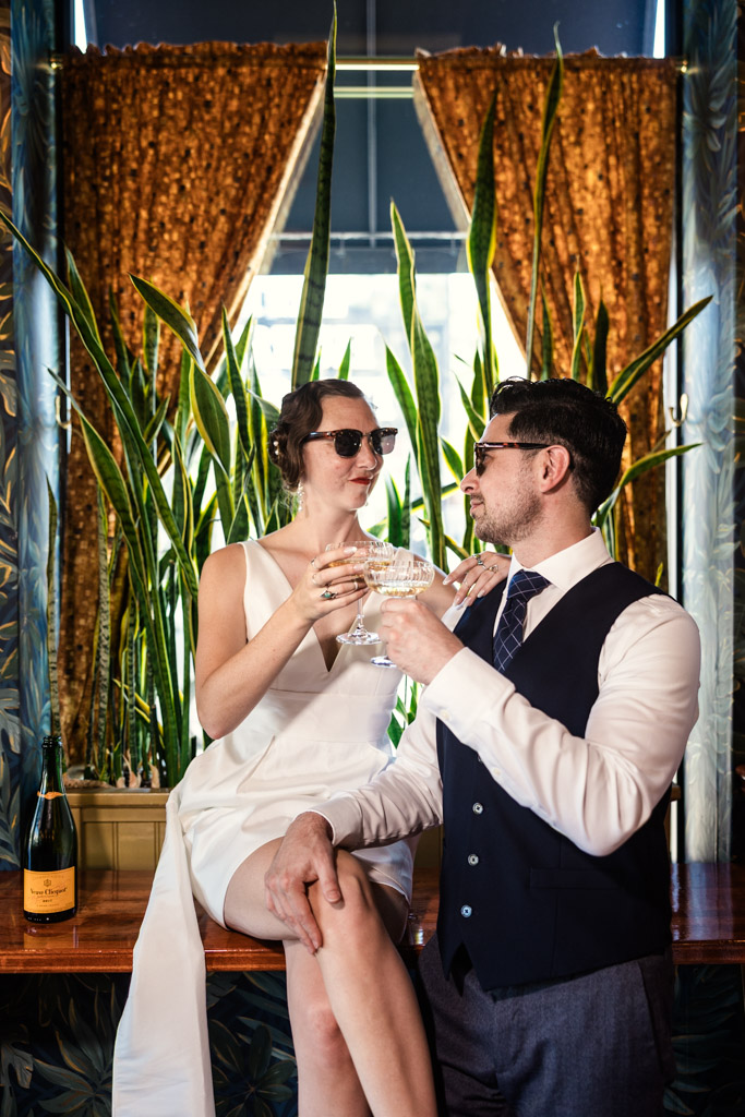 Stylish couple in formal attire clink glasses while sitting by a window adorned with plants and gold curtains behind them during Cafe Deko engagement session