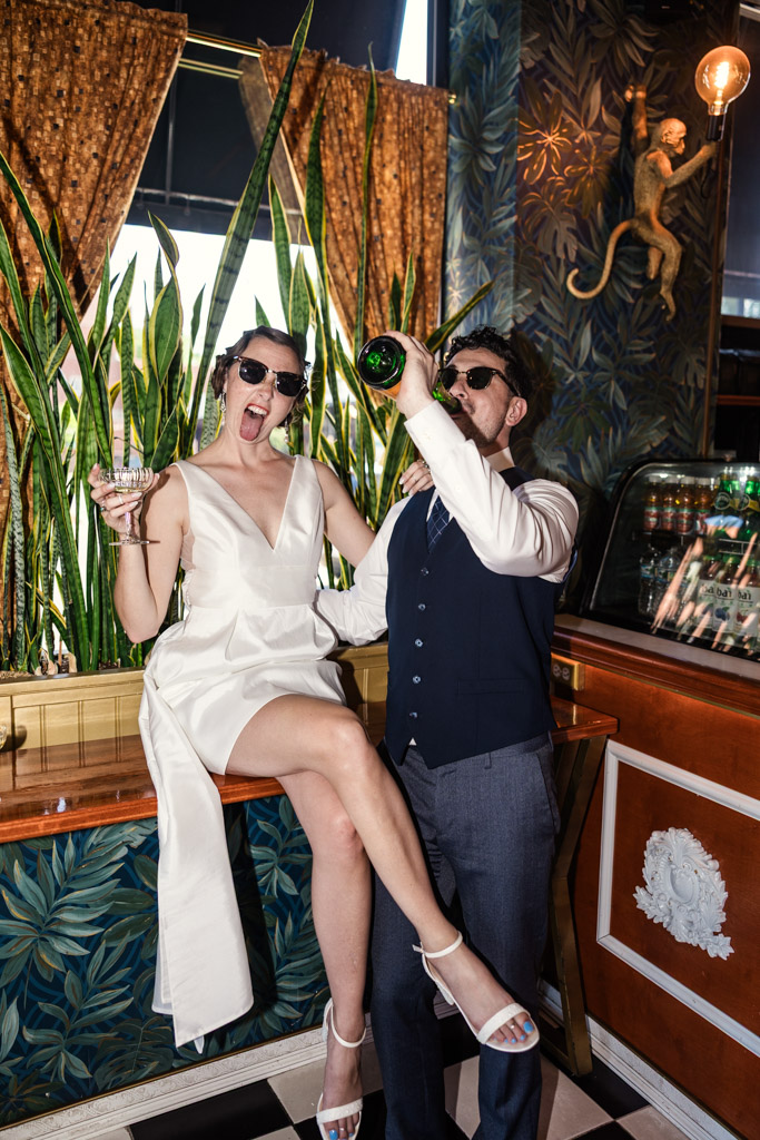 A woman in a white dress and a man in a vest celebrate their engagement with champagne in a stylish, plant-filled room at Cafe Deko in Chicago