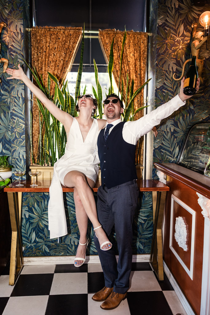 Joyful bride and groom in sunglasses celebrate with their arms raised, with plants and vintage decor behind them during Cafe Deko engagement session