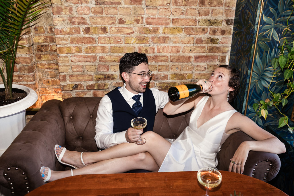 Engaged couple sits on a couch at their cocktail bar engagement; one drinks champagne from the bottle while the other laughs, holding a glass