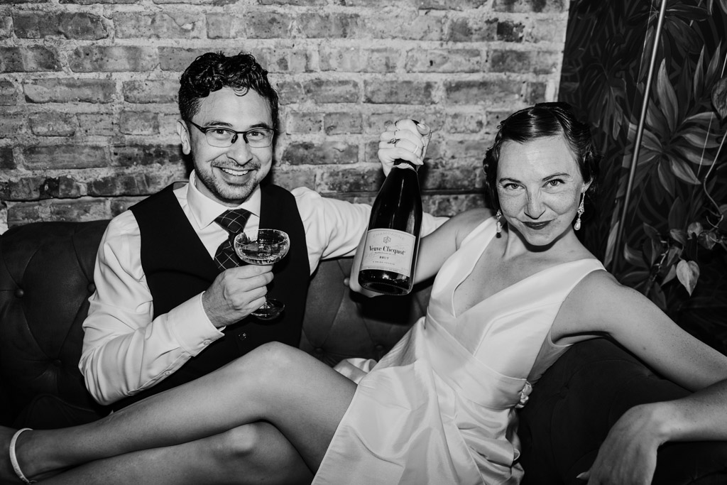 Black and white photo of smiling couple on a couch, dressed in formal attire, celebrating their cocktail bar engagement with a champagne bottle and glass