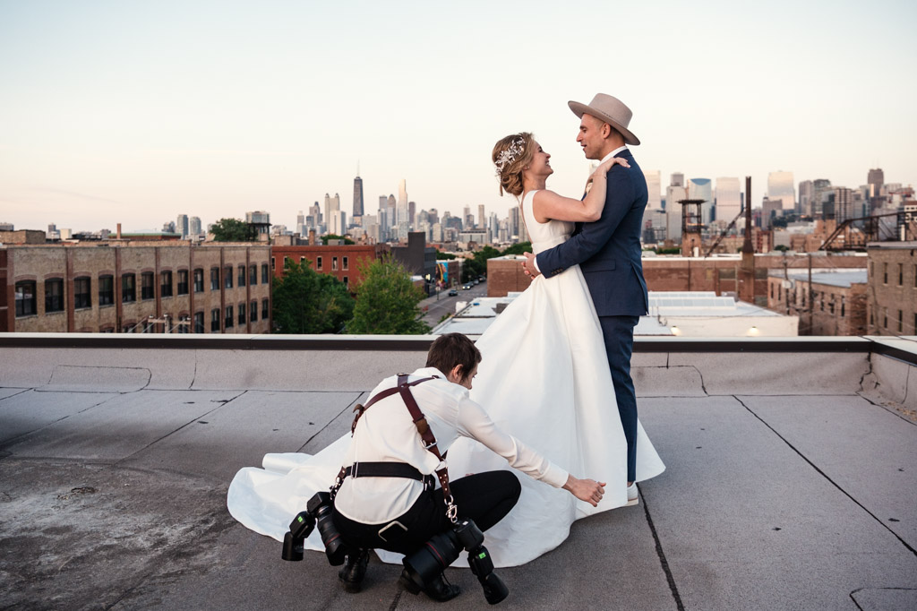 A documentary wedding photographer arranges a bride's dress as she poses with her groom on a city rooftop at sunset.