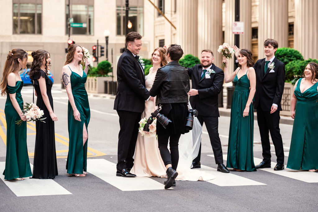 A wedding party poses and laughs on a city street as a documentary wedding photographer directs them on a crosswalk.