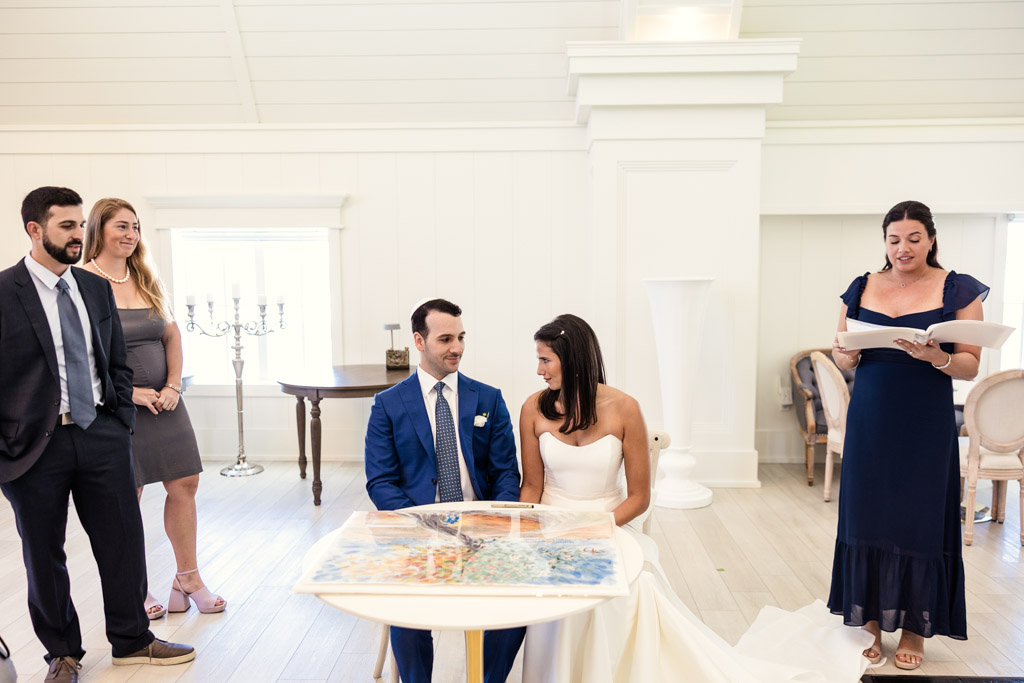 Bride and groom sit at a table before signing their ketubah inside a bright room at Être Farms