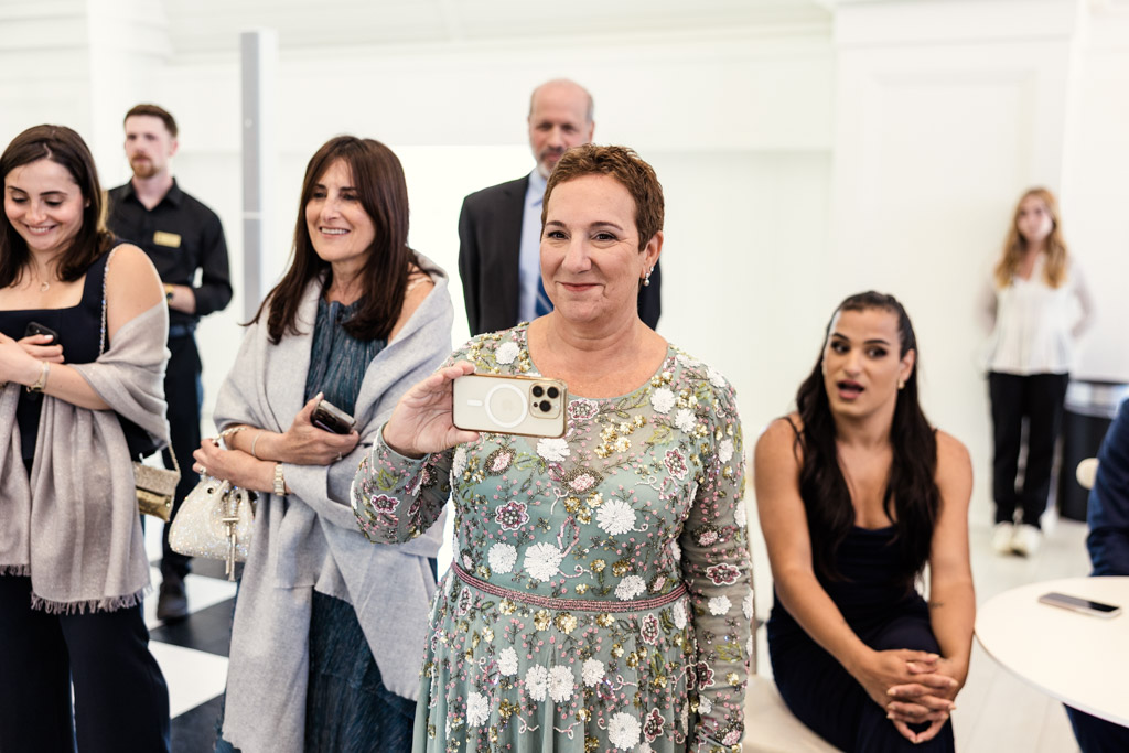Bride's mother in a floral dress smiles and holds a phone recording the signing of the ketubah during Être Farms wedding celebration