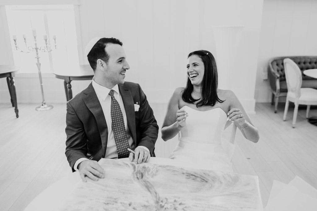 Black and white photo of bride and groom sitting together, smiling and laughing at a table as they sign the ketubah during their Être Farms wedding celebration