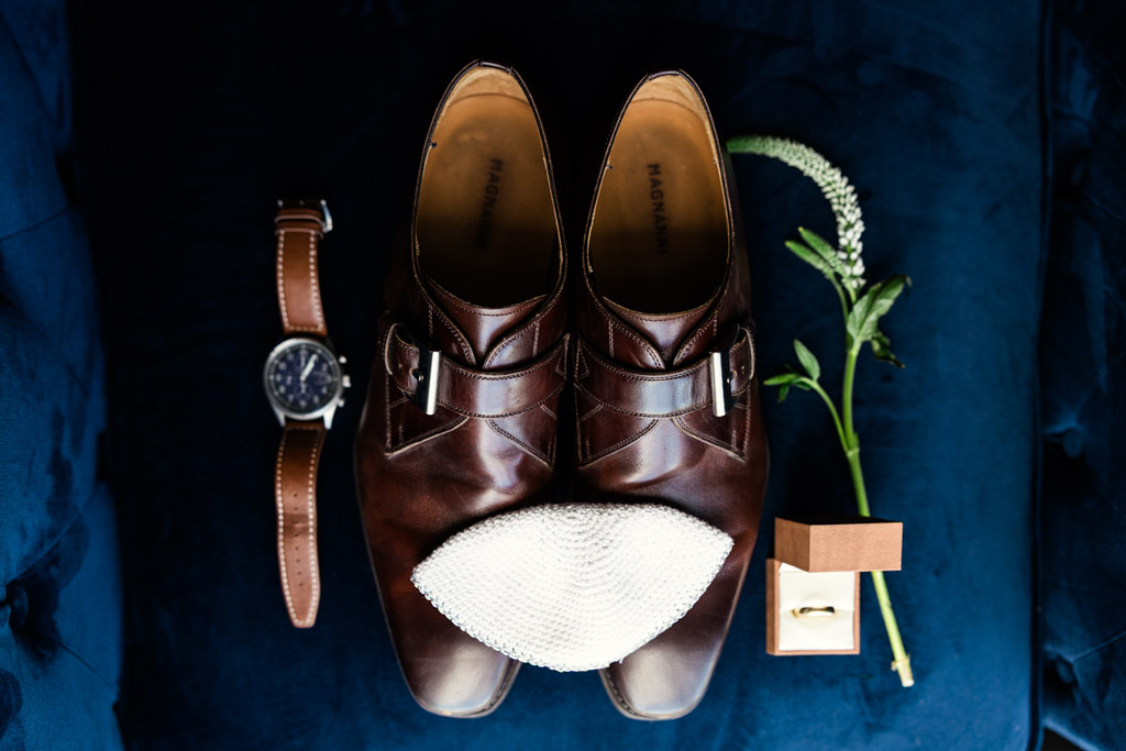 Brown dress shoes, a watch, white kippah, boutonniere, and ring box arranged on a dark blue surface at Être Farms