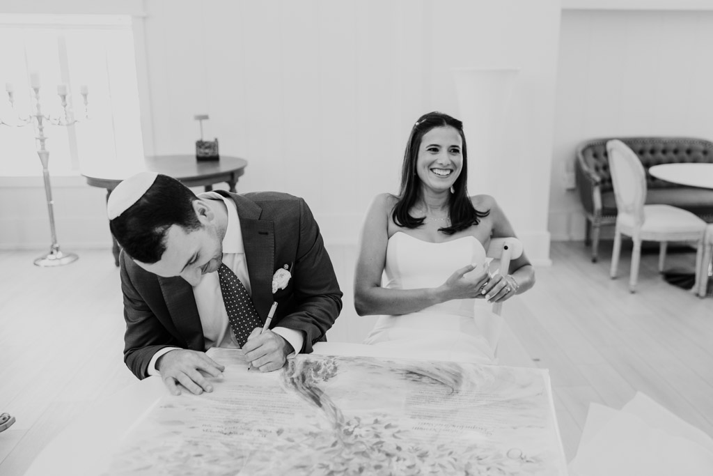 Black and white photo of groom signing the ketubah while the smiling bride sits beside him in a bright room at Être Farms