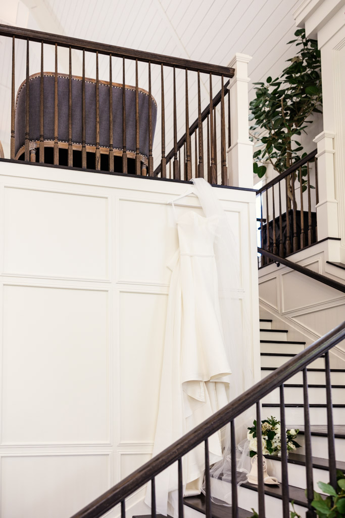 A white wedding dress and veil hang on a staircase railing in the bright interior of Être Farms
