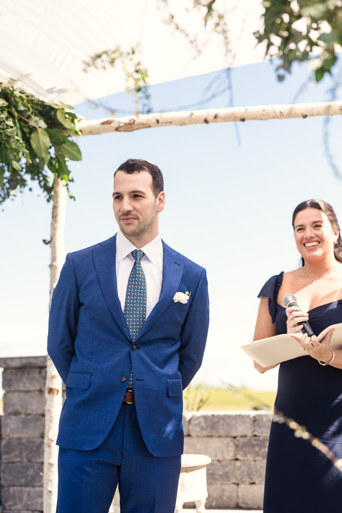 Groom in a blue suit watches teh bride walk down the aisle during their spring Être Farms outdoor wedding ceremony