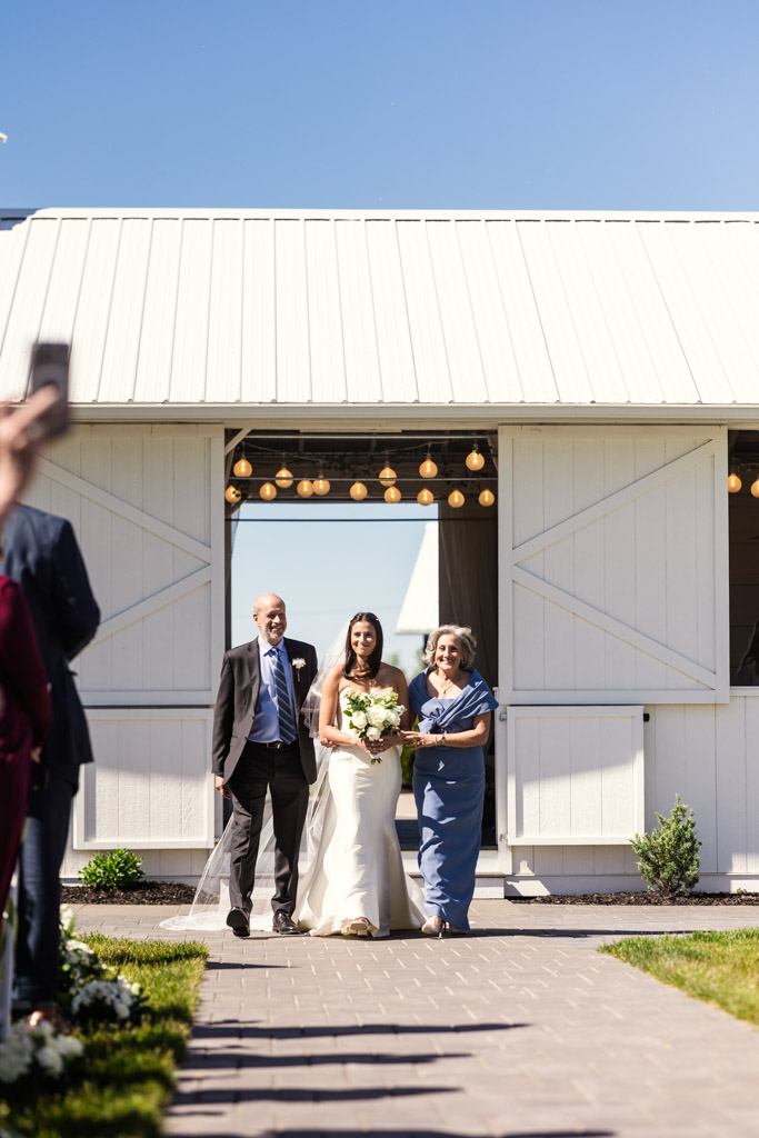 Bride in a white dress walks down the aisle with her parents outside the a white barn at Être Farms