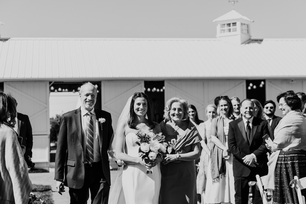 Black and white photo of bride walking down the aisle with her parents, smiling and surrounded by guests at her outdoor Être Farms wedding ceremony