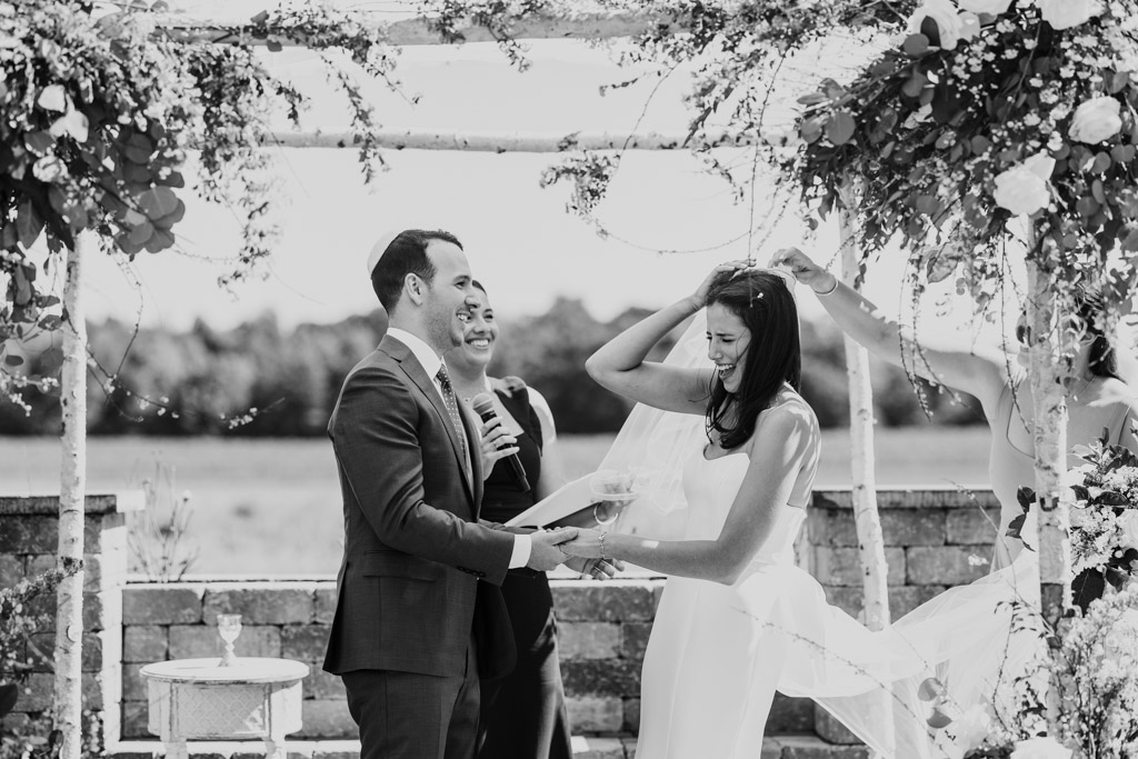 Black and white photo of bride and groom laughing as the wind catches the bride's veil during an outdoor Être Farms wedding ceremony