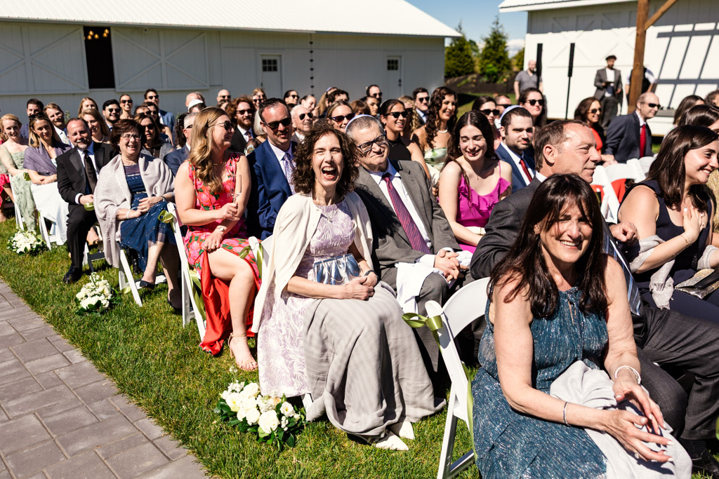 Wedding guests seated outdoors at Être Farms ceremony, smiling and laughing, with white flowers lining the aisle on a sunny day