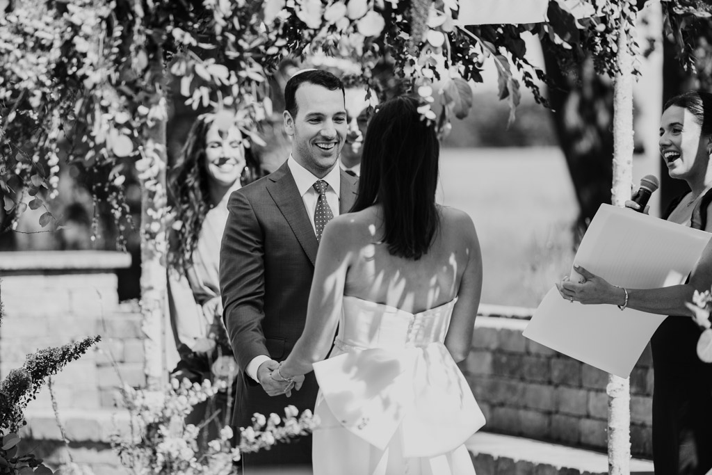 Black and white photo of groom smiling and holding bride's hands during an outdoor wedding ceremony at Être Farms