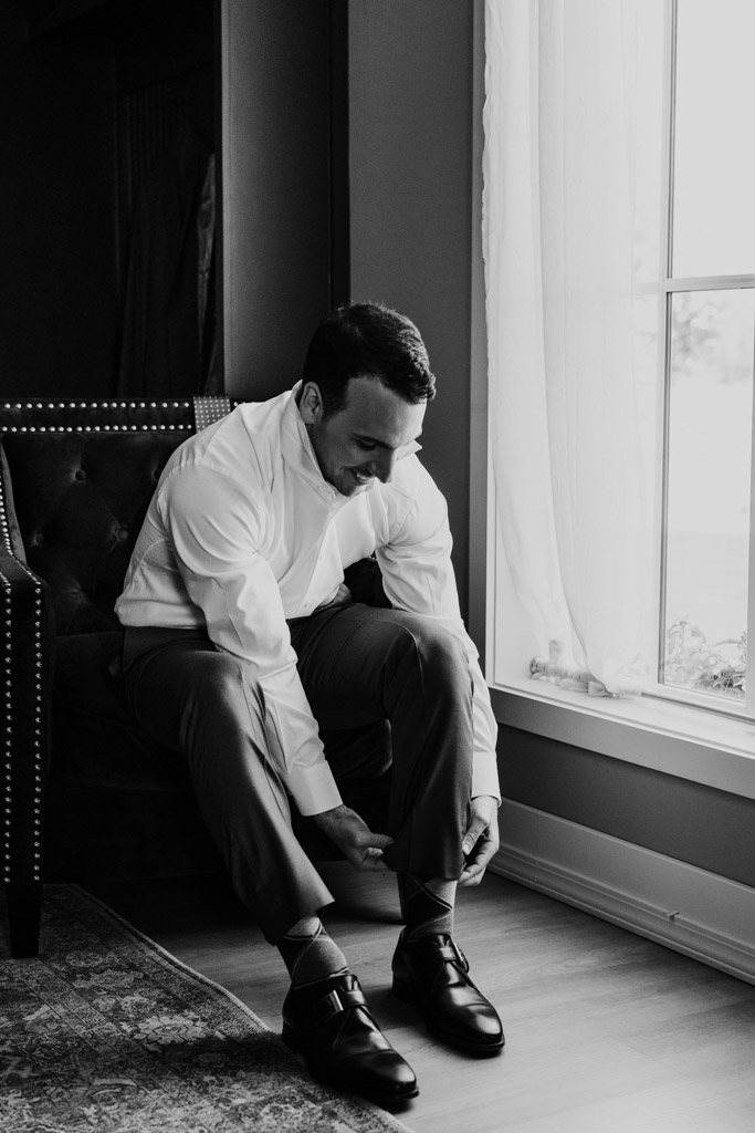 Black and white photo of groom sitting by a window, adjusting his pant leg and smiling while getting ready at Être Farms
